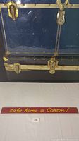 Photo showing vintage rectangular red sign with yellow cursive writing 'take home a Carton!' placed on white surface in front of a black trunk with brass hardware.