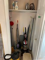 Closet view showing two upright vacuums, ironing board, two irons on shelf, broom, mop, boot mat, and step stool