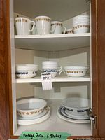 Three shelves inside a cabinet containing stacked Corelle dishes in two color patterns: orange floral and blue floral. Includes mugs, cups, bowls, and plates.