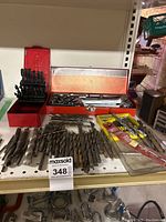 Wide view of the lot showing drill bits, socket wrench set, and wrenches in a red metal case on a white shelf.