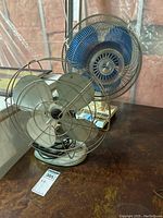 Two fans side by side on a table, a retro metal fan with large blades in front, and a blue multi-speed fan behind.