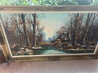 Framed oil painting of a cabin in the woods by a water body, detailing the whole artwork and frame shape and condition.