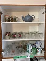 Wide view of three shelves with two large Pyrex mugs, teapot, multiple shot glasses, clear glass mugs, and colorful ceramic mugs.