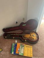 Archtop guitar in open hard case with five sheet music books positioned next to it on floor.
