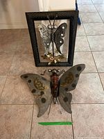 Both metal butterfly decor pieces displayed on tile floor: framed butterfly in black frame with floral metal accents and free-standing butterfly sculpture with glass bottle body.