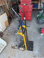 Side angle of the metal handcart showing the yellow frame, foldable base plate, wheels, and black handles.