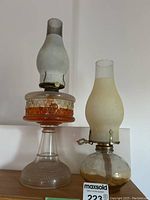 Two vintage glass oil lamps displayed with the taller amber and clear patterned glass lamp on the left and smaller round clear glass lamp on the right. Both have frosted glass chimneys and metal fixtures.
