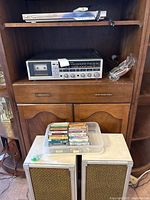 Photo showing the full lot in a wooden cabinet: DVD player on top shelf, stereo system below, plastic tub with cassette tapes on a white marble cabinet, and two speakers on floor.