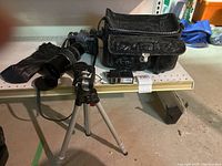 Wide view of the lot showing the camera on the right with long attached lens, black cloth bag, compact tripod with legs deployed, and separate lens caps and smaller lens on shelf.