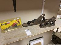 Three wood planers on a shelf: two used metal planers with wooden handles and front knobs, and one new Stanley hand planer in sealed packaging with red and black handle.