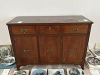 Front-facing photo of wooden sideboard showing three lower cabinets and two drawers above, with brass handles and decorative carvings. Visible marks and water damage on top surface.