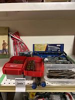 Various drill bits in Milwaukee cases and loose, a blue socket set with ratchets and extensions, and red pouch with long sockets arranged on a shelf