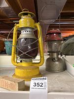 Two vintage lanterns on a white surface; foreground large yellow Beacon lantern with cracked purple glass globe, background smaller silver and red Coleman lantern