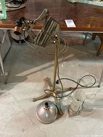 Full view of the heavy vintage floor lamp stand with its accordion arm, tripod base, exposed wiring and a metal shade detached and placed on the floor.