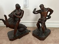 Front view of both wooden sculptures, showing a man wrestling a lion on the left and a man wrestling a snake on the right, placed on carpeted floor.