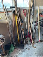 Photo showing a variety of yard tools including multiple rakes, shovels, hoes, and a pitchfork arranged against a wall in a garage or shed environment.