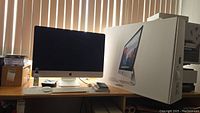 Front view of powered-off 27″ iMac on desk with Apple Wireless Keyboard and Magic Mouse in front, original box at right