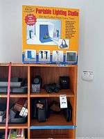 Front view of Digital Portable Lighting Studio box on top shelf above wooden shelving containing Nikon binoculars with case, Trilyte binoculars, and various carrying cases.