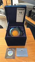 Paperweight displayed inside navy blue velvet box with interior satin lining, including the Glass Eye Studio Celestial Series certificate and information card in front.