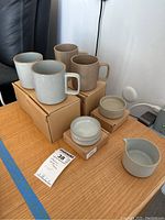 Photo showing two light grey porcelain mugs, two beige porcelain mugs, small round lids/coasters, and a creamer, all on a wooden surface with some original cardboard boxes.