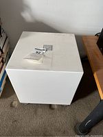 White cube-shaped subwoofer with four square metal feet on carpet, top view of flat surface