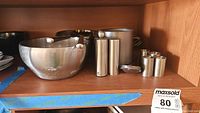 Shelf holding silvertone stacking mug, Peugeot salt and pepper shakers, stainless steel bowls and other kitchenware items