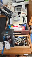 Photo showing multiple stationery items on a desk, including pens, markers, tape rolls, scissors in a wooden box, and boxed X-ACTO sharpener.