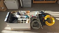 Box with assorted electronics including surge protector boxes, cables, and plugs