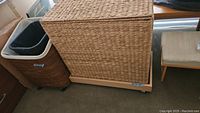 Front and side view of the wicker hamper showing woven texture, lid closed, beige laundry bag visible inside two compartments, and caster wheels under wooden base.
