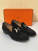 Pair of black suede Hermès loafers positioned in front of an orange Hermès box on a white surface