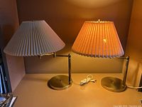 Two articulating brass lamps side by side with pleated shades, one lit amber and one white, cords visible