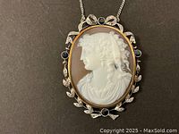 Front view of oval shell cameo pendant showing carved profile, gold bezel, diamond-set leaf motifs and four blue sapphires