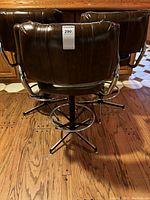 Back view of three brown swivel bar stools showing high backs and armrests, metal bases, and wooden floor.