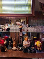 Photo showing seven Pinwheel crystal highball glasses and various glass tumblers arranged on mirrored shelf. Below are plastic M&M items, beer cozies, and Waring martini shaker.