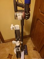 Dyson cordless vacuum assembled on white stand with hanging brackets and charging dock, showing main vacuum unit and handle with trigger and clear dustbin.