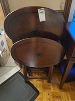 Four oval wooden folding tables stacked with visible compass star inlay designs on top. Tables are placed next to a leather chair and other furniture.