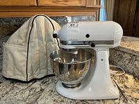 White KitchenAid stand mixer assembled with stainless steel bowl on countertop alongside quilted cover