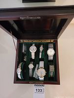 Photo showing six ladies watches arranged in a wooden display case with green velvet compartments, various styles and colors visible.