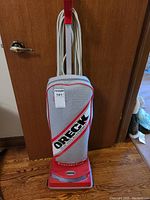 Full front view of an Oreck Cloverdale Hypo-Allergenic upright vacuum cleaner with wrapped cord, standing against a wooden door.
