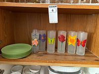 Shelf with 8 green luncheon plates stacked and 11 frosted glass tumblers with painted flowers and butterflies