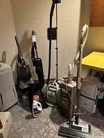 Wide shot showing group of seven vacuum cleaners in various colors and styles plus bottle of vacuum fluid on tiled floor