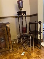 Wide view showing tall twisted-leg plant stand with candle holder, shorter three-shelf plant stand, red vase inside tall stand, and metal magazine rack in front.