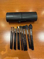 Set of 11 Sigma makeup brushes aligned in size order on wooden surface, with a black cylindrical brush holder case placed behind them.