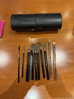 Twelve Sigma brushes displayed in a row alongside a closed black cylindrical brush holder case on a wooden surface.