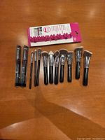 Full set of eleven Sigma brushes laid out with the pink wall suction brush holder behind them on wooden surface.