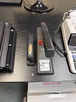 Top view showing both black staplers side by side on a dark wooden desk, including the mega stapler with red adjustment part and the smaller normal stapler with gold oval badge.