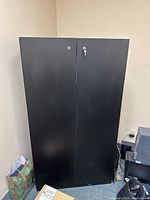 Front view of the tall black plywood cabinet with closed double doors and visible lock with key.