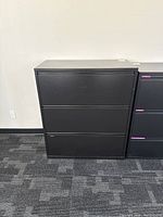 Front view of black filing cabinet showing three horizontal drawers and flat top surface.