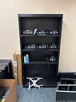 Front view of black wooden bookshelf with several electronic calculators and a drone placed in front and around it, along with some boxed items on the left side.