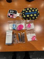 Makeup bag, hand mirror, various makeup sponges, boxed eyelash curler, tweezers, and makeup brushes displayed on wood surface from overhead angle.